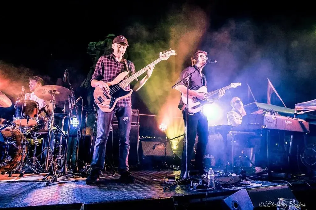 Drummer, bassist, guitarist, and keyboard player performing under colorful stage lights